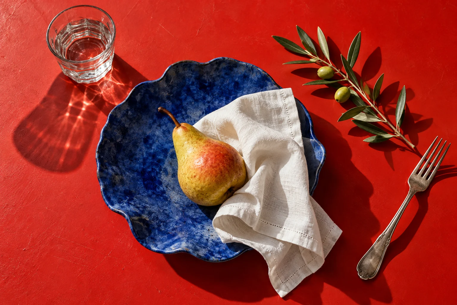 A pear on a blue ceramic plate with linen, glass and olive branch on a red table