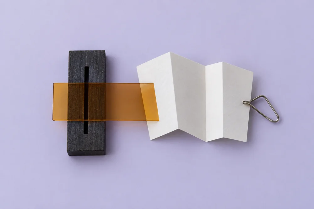 Generated overhead material study: charcoal slotted block, amber strip, folded white paper and bent silver wire on lilac.