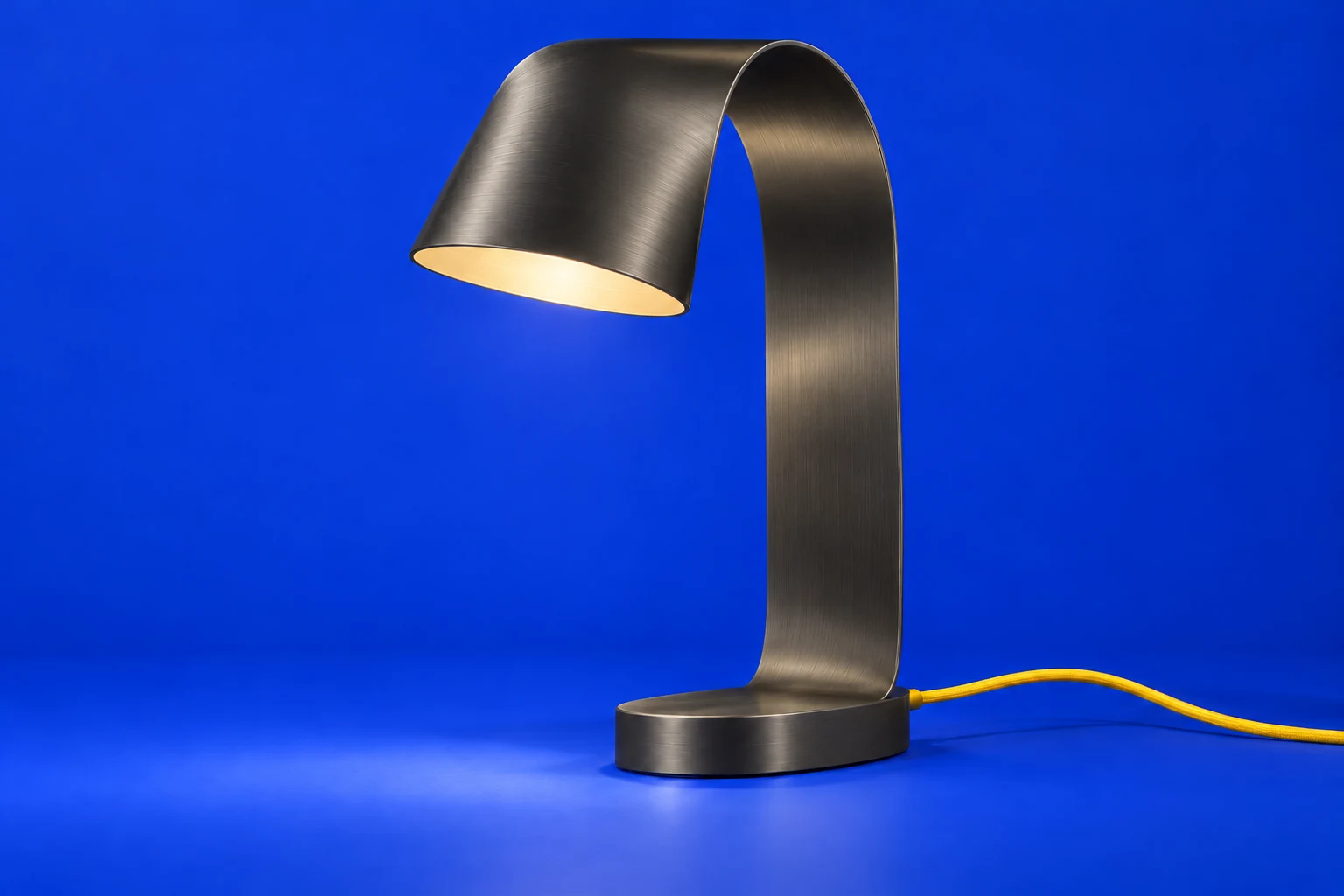 Rill 01, a sculptural ribbon-shaped aluminum lamp with a yellow cable on a cobalt stage.