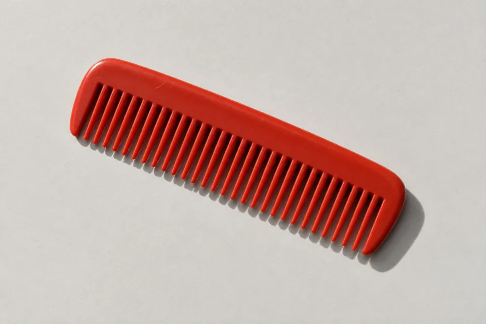 A red pocket comb with a single row of teeth lying diagonally on pale gray paper.