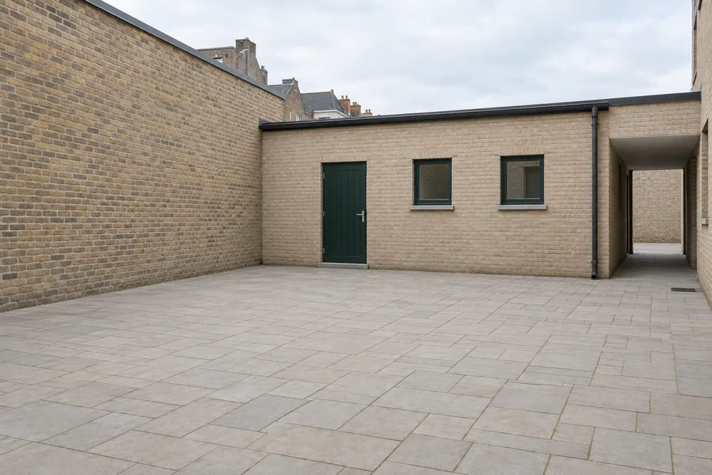 Fictional existing courtyard: bare paving, brick walls, green door, two windows and a passage at right.