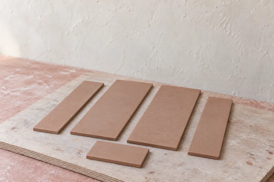 Conceptual flat clay-colored wall and base pieces for the tall rectangular vessel, arranged on a pale work board.