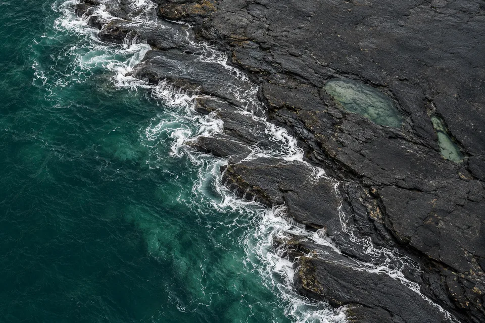Near-vertical generated view of dark low basalt shelf meeting jade water and pale foam, with two small rock pools