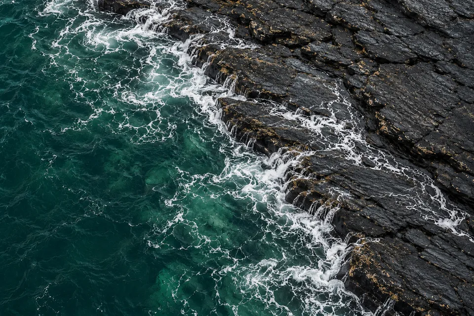 Generated close overhead view of thin pale foam meeting layered dark rock beside jade seawater
