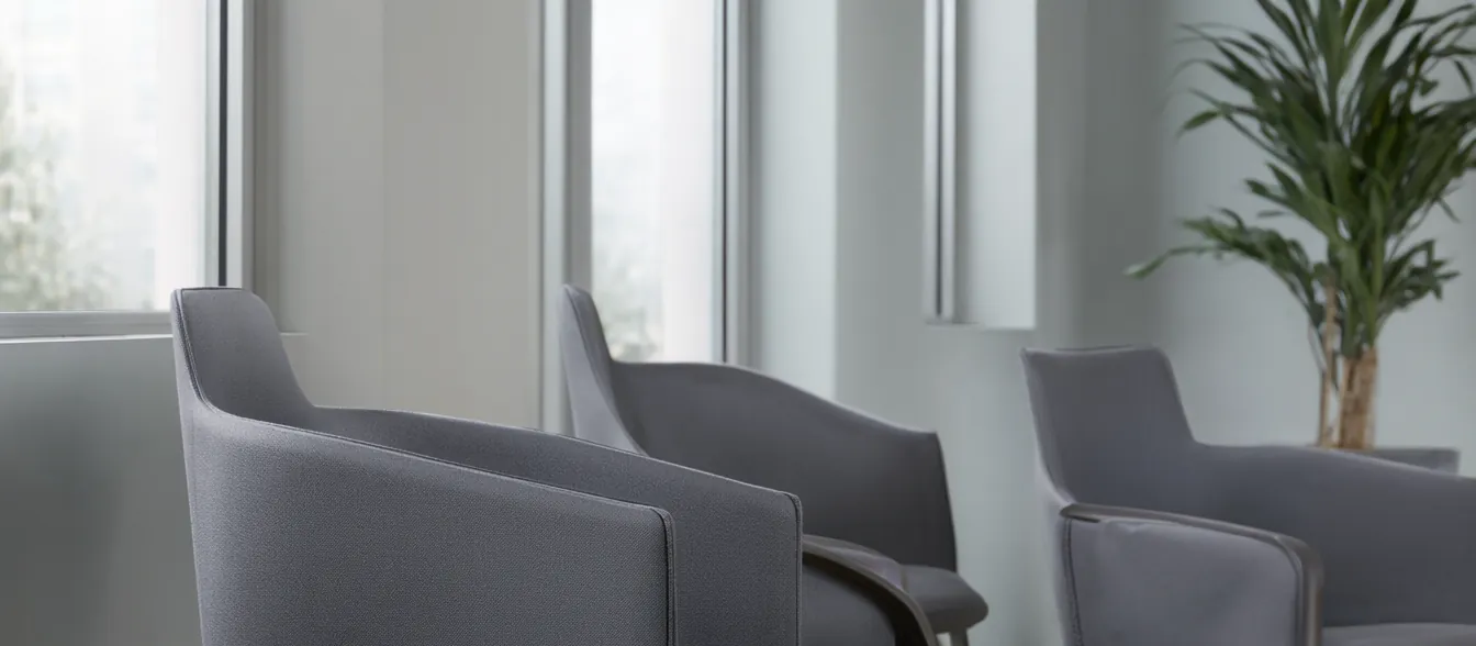 A calm waiting room with grey chairs, a potted plant and daylight from tall windows.
