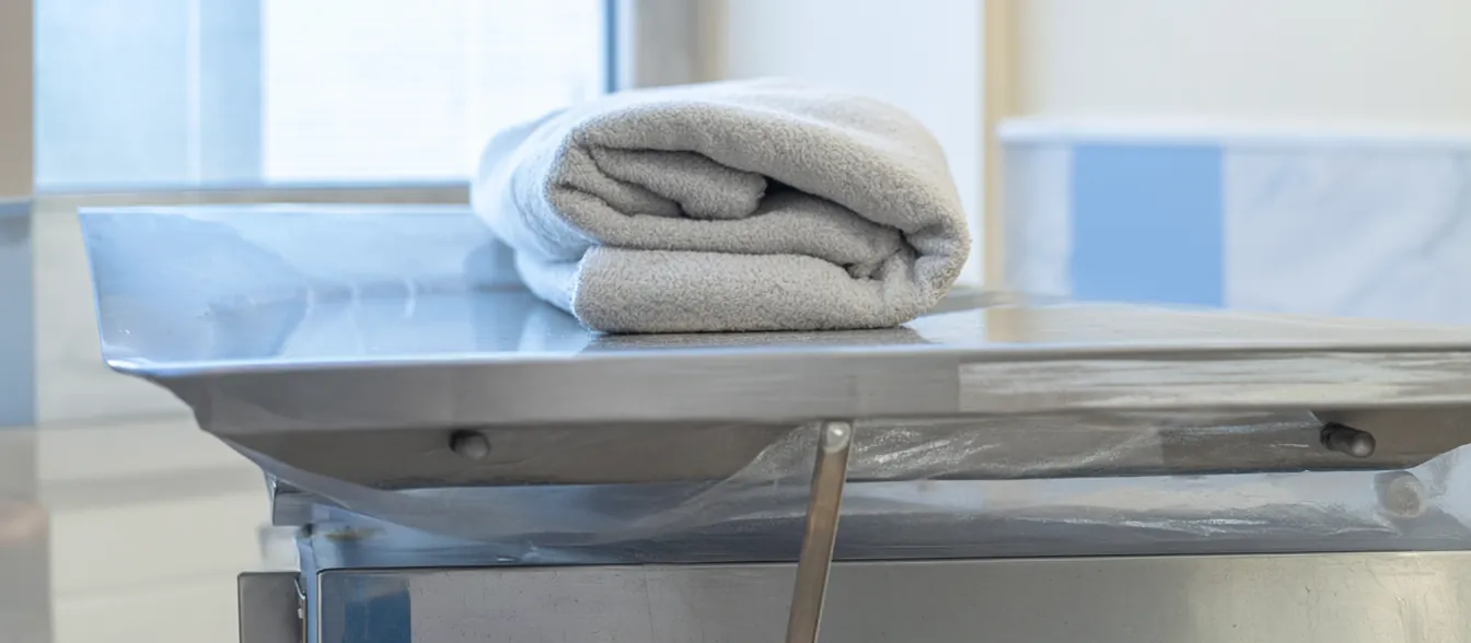 A bright exam room with a steel table, a folded blanket and a window with natural light.