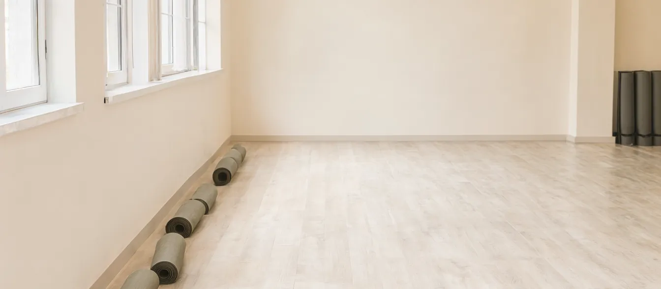 A bright studio room with pale wood floors, rolled mats and tall windows.